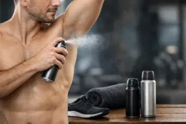A shirtless man applies deodorant spray to his armpit. Nearby are two spray cans, a rolled towel, and a running shoe, conveying a gym setting and freshness.