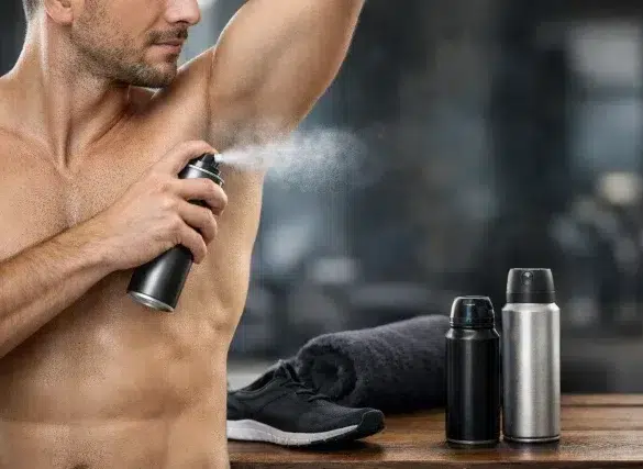 A shirtless man applies deodorant spray to his armpit. Nearby are two spray cans, a rolled towel, and a running shoe, conveying a gym setting and freshness.