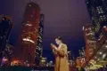 Woman in a tan coat smiles at her smartphone against a backdrop of brightly lit skyscrapers at night, conveying a sense of connectivity and urban energy.