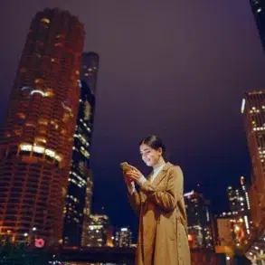 Woman in a tan coat smiles at her smartphone against a backdrop of brightly lit skyscrapers at night, conveying a sense of connectivity and urban energy.