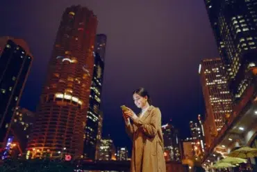 Woman in a tan coat smiles at her smartphone against a backdrop of brightly lit skyscrapers at night, conveying a sense of connectivity and urban energy.