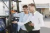 Two men stand beside a blue tractor. One holds a key while the other points at it, smiling, with a green notebook in hand, suggesting a transaction or lesson.