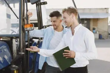 Two men stand beside a blue tractor. One holds a key while the other points at it, smiling, with a green notebook in hand, suggesting a transaction or lesson.