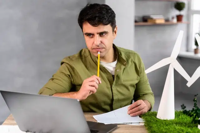 Man in green shirt at desk, holding pencil thoughtfully, looking at laptop. Paper wind turbine model on grass mat nearby. Focused and contemplative mood.