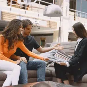 A couple sits on a sofa, attentively examining fabric samples with a saleswoman in a showroom. The setting is bright and contemporary.