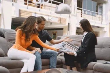 A couple sits on a sofa, attentively examining fabric samples with a saleswoman in a showroom. The setting is bright and contemporary.