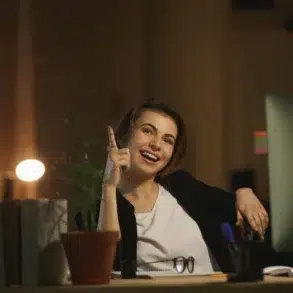 Cheerful woman in a dimly lit office, pointing upward with an idea, seated at a desk with a computer, potted plant, books, and stationery. Warm, motivated atmosphere.