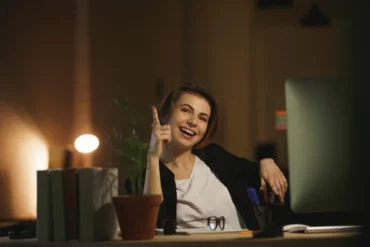 Cheerful woman in a dimly lit office, pointing upward with an idea, seated at a desk with a computer, potted plant, books, and stationery. Warm, motivated atmosphere.