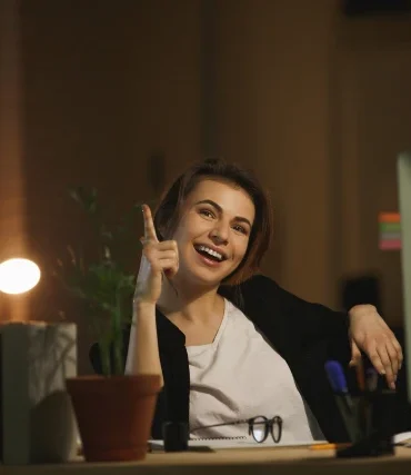Cheerful woman in a dimly lit office, pointing upward with an idea, seated at a desk with a computer, potted plant, books, and stationery. Warm, motivated atmosphere.