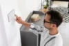 Man in glasses touches a wall-mounted smart device, in a modern, minimalist room with shelves and a rug, conveying focus and modern technology.