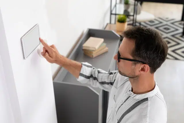 Man in glasses touches a wall-mounted smart device, in a modern, minimalist room with shelves and a rug, conveying focus and modern technology.