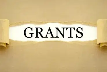 The word 'Grants' is revealed by peeling back a layer of beige paper, symbolizing discovery and opportunity. The background is a smooth, muted gold.