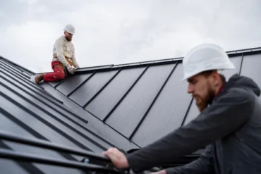 Two construction workers on a metal roof, wearing white hard hats and work attire. One focuses on installing roofing material; the other secures a ladder, under a cloudy sky.