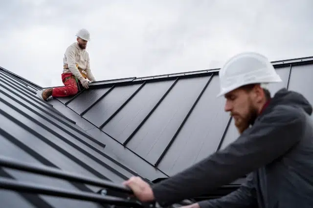 Two construction workers on a metal roof, wearing white hard hats and work attire. One focuses on installing roofing material; the other secures a ladder, under a cloudy sky.
