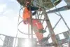 Worker in orange safety gear climbs a rusted metal ladder on an industrial structure. Sunlight streams through, contrasting with the blue sky, conveying determination.