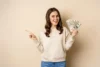 Smiling woman in a beige sweater holds a fan of dollar bills, pointing to the side against a plain background, conveying excitement and success.