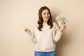 Smiling woman in a beige sweater holds a fan of dollar bills, pointing to the side against a plain background, conveying excitement and success.