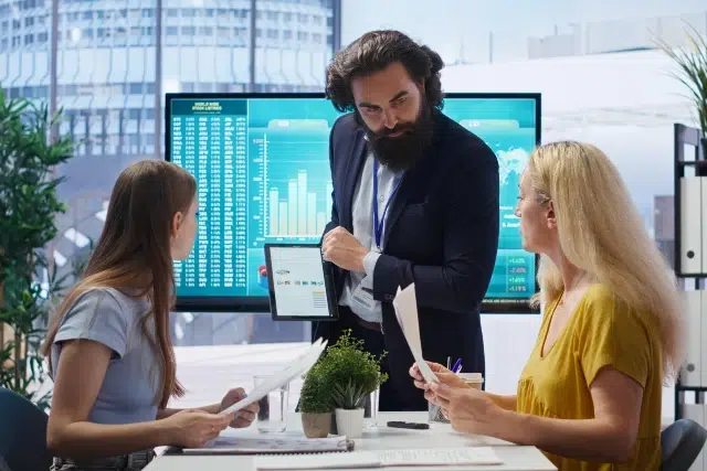 A bearded man in a suit presents data on a tablet to two women in an office. They're discussing charts on screens, creating a focused, analytical atmosphere.