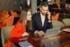 A man and woman in professional attire sit at a wooden table in a cafe, smiling while looking at a laptop. She wears a red blazer, and he's on the phone.
