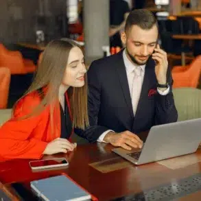 A man and woman in professional attire sit at a wooden table in a cafe, smiling while looking at a laptop. She wears a red blazer, and he's on the phone.
