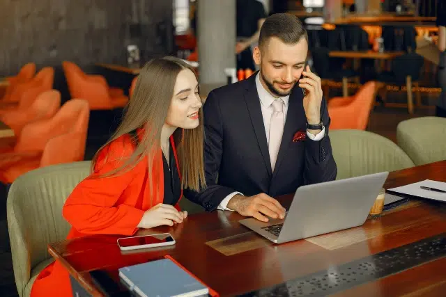 A man and woman in professional attire sit at a wooden table in a cafe, smiling while looking at a laptop. She wears a red blazer, and he's on the phone.