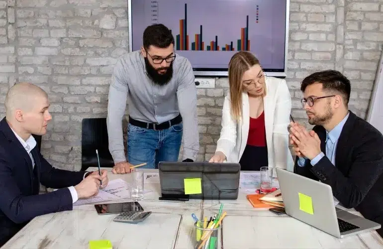 A team of four people in an office discusses charts displayed on a laptop and a screen. The mood is focused, teamwork emphasized, brick wall behind.