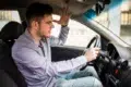 A frustrated driver in a parked car gestures with his hand, expressing irritation. He wears a light shirt, with a tense expression on his face.