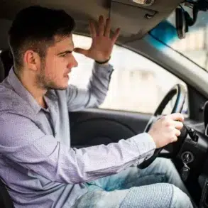 A frustrated driver in a parked car gestures with his hand, expressing irritation. He wears a light shirt, with a tense expression on his face.