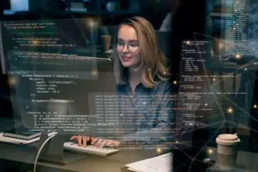 A woman with glasses smiles while working on a computer in a dimly lit office. Code and digital graphics are overlaid, conveying a tech-focused atmosphere.