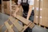 Worker in overalls handling a wooden pallet in a warehouse filled with stacked cardboard boxes, suggesting a busy, organized environment.
