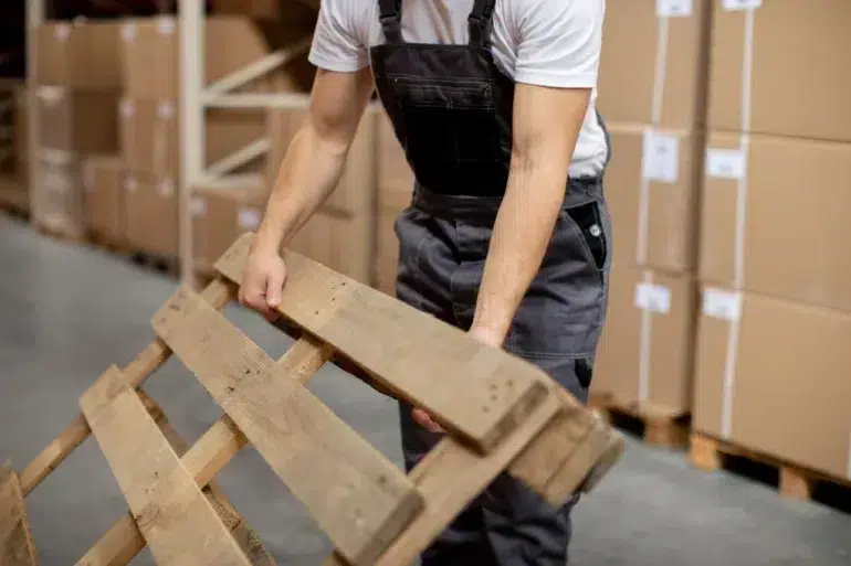 Worker in overalls handling a wooden pallet in a warehouse filled with stacked cardboard boxes, suggesting a busy, organized environment.