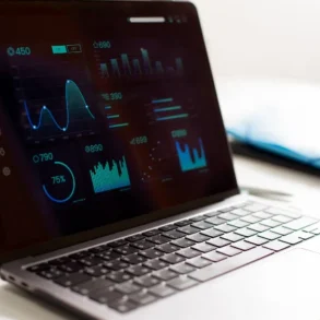 A laptop displays various digital graphs and charts with glowing blue and green data on a dark screen, suggesting data analysis or business analytics.