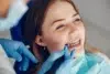 Young woman with braces smiles during a dental check-up. A dentist's hand in blue gloves uses a dental tool. The atmosphere is calm and professional.