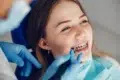 Young woman with braces smiles during a dental check-up. A dentist's hand in blue gloves uses a dental tool. The atmosphere is calm and professional.