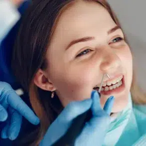 Young woman with braces smiles during a dental check-up. A dentist's hand in blue gloves uses a dental tool. The atmosphere is calm and professional.