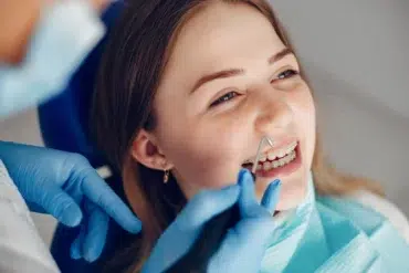 Young woman with braces smiles during a dental check-up. A dentist's hand in blue gloves uses a dental tool. The atmosphere is calm and professional.