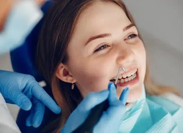 Young woman with braces smiles during a dental check-up. A dentist's hand in blue gloves uses a dental tool. The atmosphere is calm and professional.