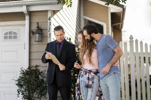 A real estate agent shows a clipboard to a couple in casual attire, standing outside a suburban house with a white picket fence, appearing thoughtful.