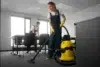 Two people in work attire clean an office. A woman vacuums with a yellow machine, while a man organizes papers. Bright, tidy room conveys diligence.
