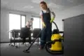 Two people in work attire clean an office. A woman vacuums with a yellow machine, while a man organizes papers. Bright, tidy room conveys diligence.