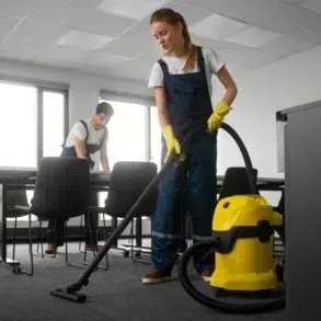 Two people in work attire clean an office. A woman vacuums with a yellow machine, while a man organizes papers. Bright, tidy room conveys diligence.
