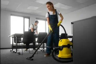 Two people in work attire clean an office. A woman vacuums with a yellow machine, while a man organizes papers. Bright, tidy room conveys diligence.
