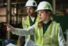 Two construction workers in hard hats and safety vests are in a factory setting. The woman in front points assertively, holding blueprints, indicating focus and direction.