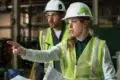 Two construction workers in hard hats and safety vests are in a factory setting. The woman in front points assertively, holding blueprints, indicating focus and direction.