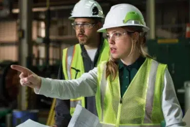 Two construction workers in hard hats and safety vests are in a factory setting. The woman in front points assertively, holding blueprints, indicating focus and direction.