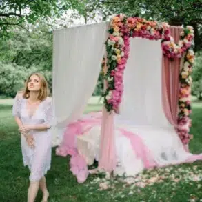 A woman in a white lace dress stands in a lush green garden near a decorated canopy bed adorned with pink and yellow flowers, exuding a whimsical, romantic vibe.