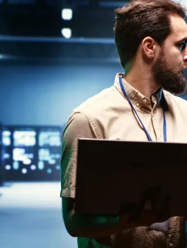 Man in a beige shirt holding a laptop in a dimly lit server room. He looks to the side, focused. Background features illuminated server racks.