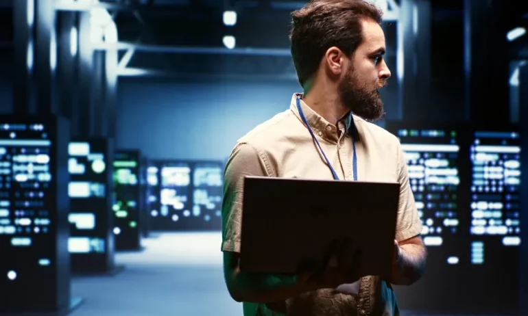 Man in a beige shirt holding a laptop in a dimly lit server room. He looks to the side, focused. Background features illuminated server racks.