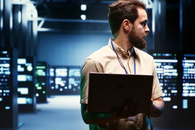 Man in a beige shirt holding a laptop in a dimly lit server room. He looks to the side, focused. Background features illuminated server racks.