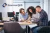 A team of three people collaborates around a desk in a bright office. Two women and a man are focused on documents, smiling. ScanmarQED logo in the corner.